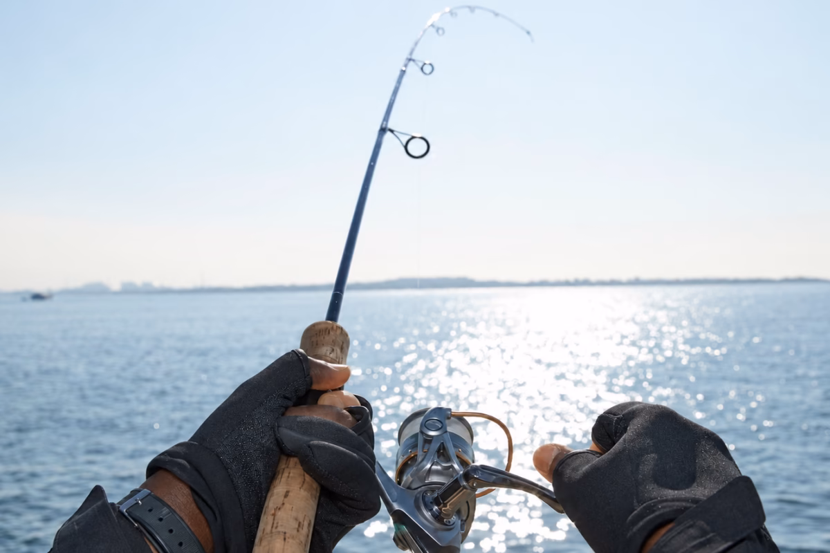 Angler's gloved hands holding a bent fishing rod over sunlit sea
